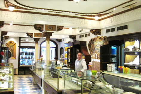Interior d'una pastisseria històrica amb vitrines plenes de dolços i un venedor al fons.