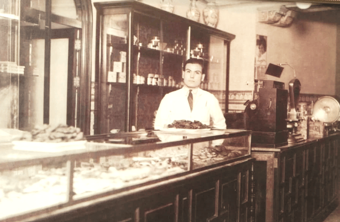 Un home jove vestit de blanc darrere d'un taulell en una botiga antiga, amb vitrines plenes de productes i una caixa registradora a la dreta.